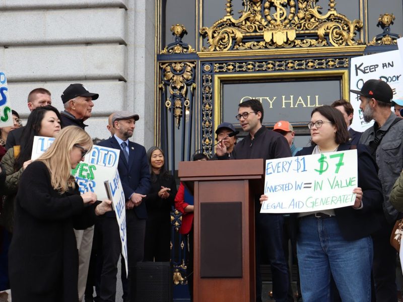A group of people gathers outside city hall holding signs in support of legal aid, while a speaker addresses the crowd from a podium.