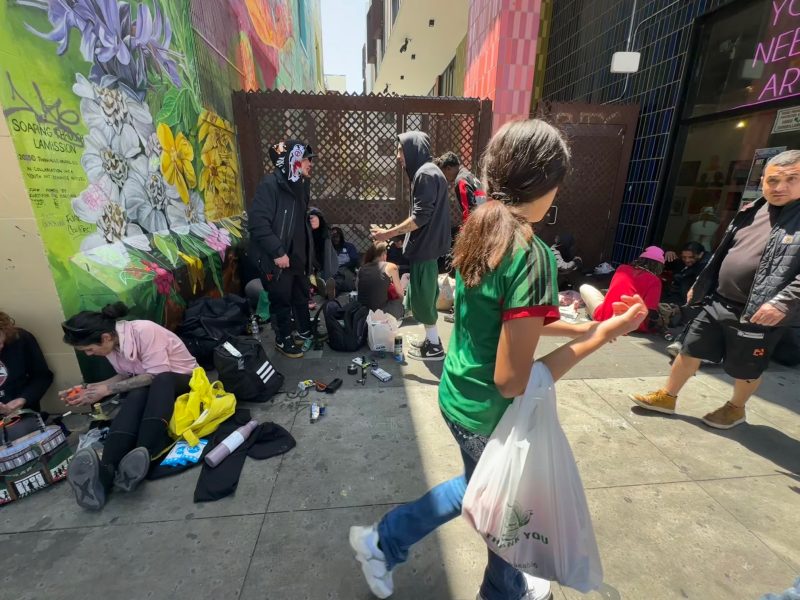A group of people sit and stand on a 16th street city sidewalk surrounded by bags and personal items; one person carries shopping bags while others interact or rest.
