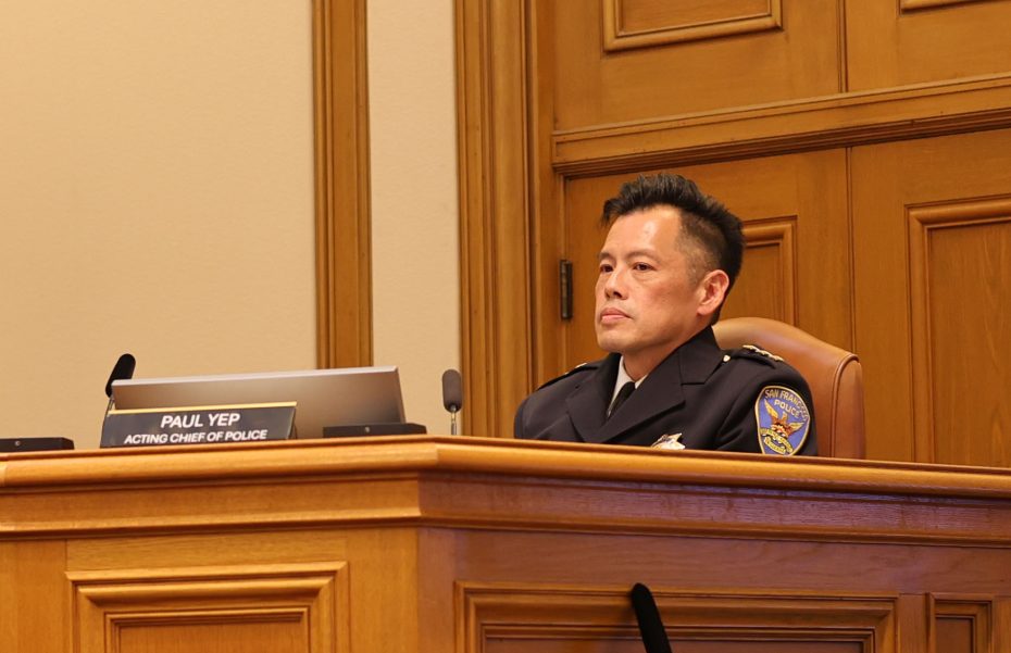 A uniformed police officer sits at a wooden desk with a nameplate reading “Paul Yep, Acting Chief of Police” in a formal police department office.