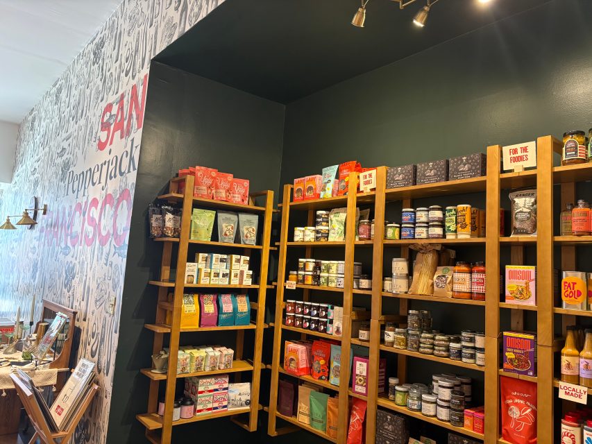 Shelves stocked with various packaged foods, jars, and boxes are displayed against a dark green wall in a specialty grocery store.
