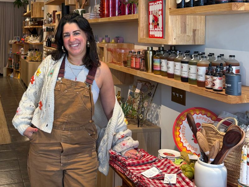 A woman wearing overalls and a sweater stands and smiles inside a well-lit shop filled with various household goods and decor items arranged on shelves and tables.