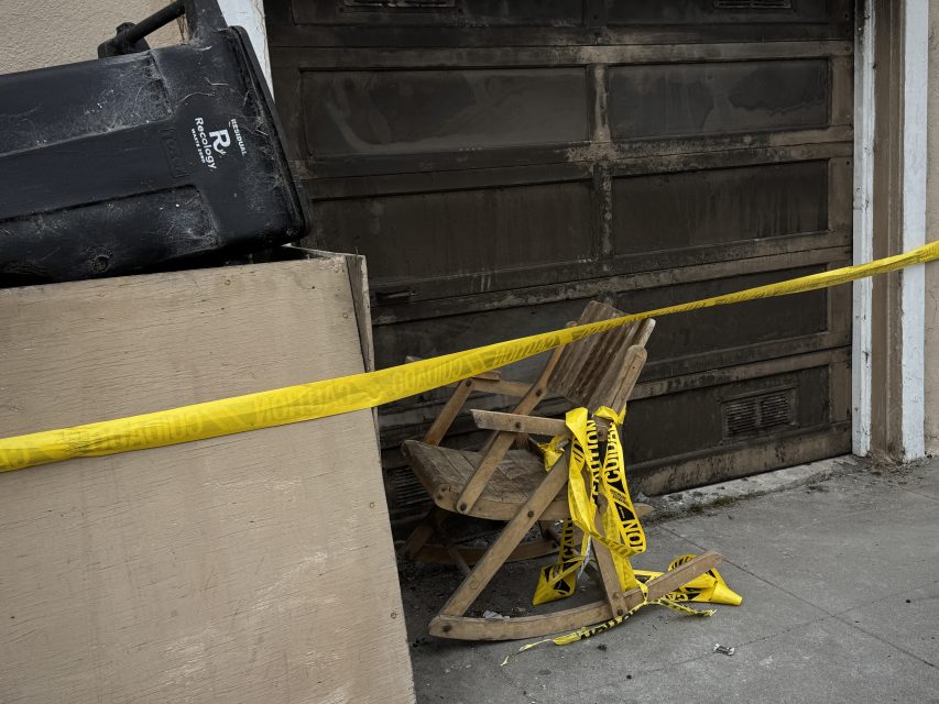 A broken wooden rocking chair lies behind yellow caution tape, next to a large trash bin and plywood panel in front of a dirty garage door.