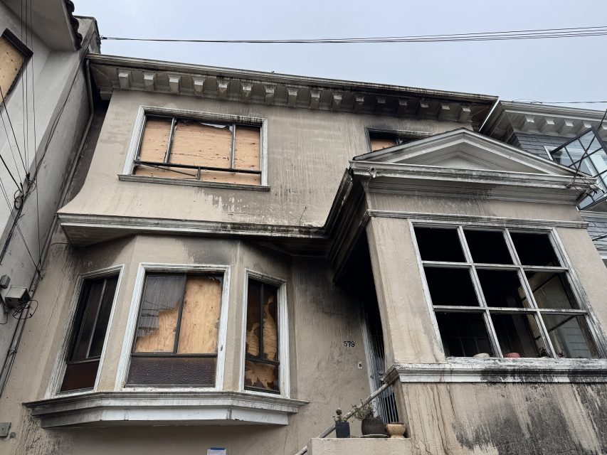 Two-story house with boarded-up and broken windows, dirty exterior walls, and visible signs of neglect.