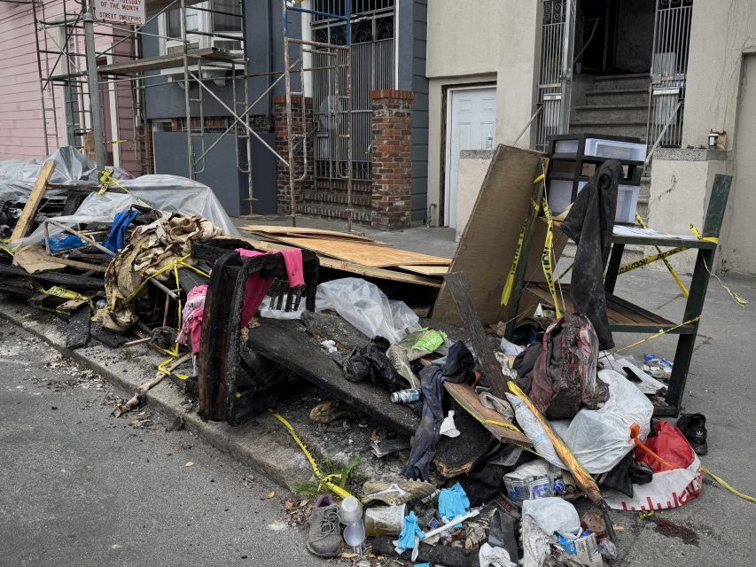 A pile of burnt debris, trash, and damaged items is stacked on a sidewalk next to a building under construction, with yellow caution tape surrounding the area.
