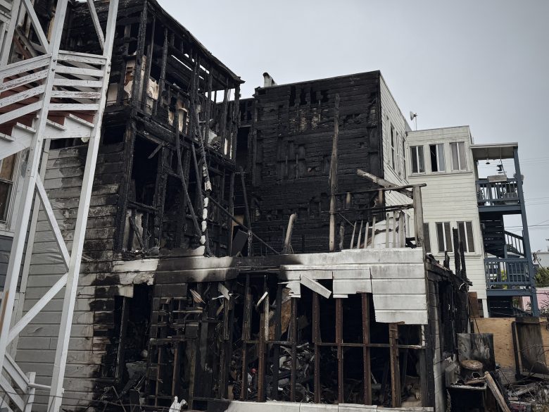 A severely burned building with charred walls and exposed framing next to undamaged neighboring structures.