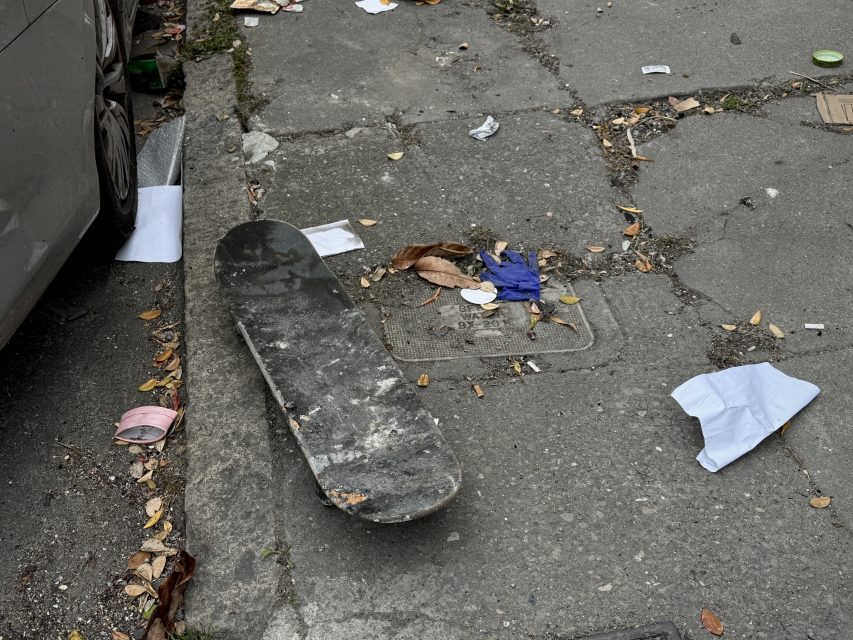 A worn skateboard lies upside down on a cracked, litter-strewn sidewalk next to a car.