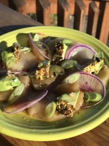 A yellow plate with thinly sliced radishes, avocado pieces, green onions, and a sprinkling of seeds or spices, arranged outdoors in sunlight.
