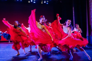 A group of dancers in red dresses perform energetically on stage under bright lighting, with flowing fabric and expressive movements.