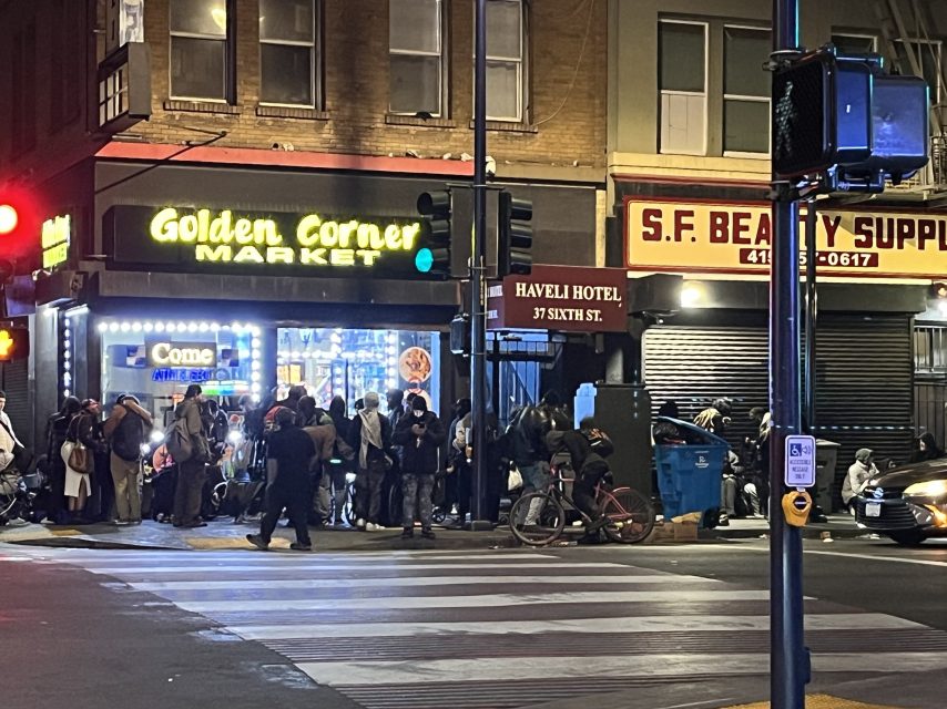 A group of people gathers outside Golden Corner Market and nearby shops at night on a city street corner with illuminated signs.