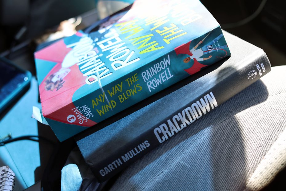 Two books stacked on a surface: "Any Way the Wind Blows" by Rainbow Rowell on top and "Crackdown" by Garth Mullins underneath.