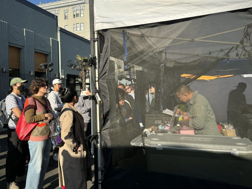 People stand in line outside a booth where a vendor prepares items behind a mesh screen at an outdoor market in daylight.