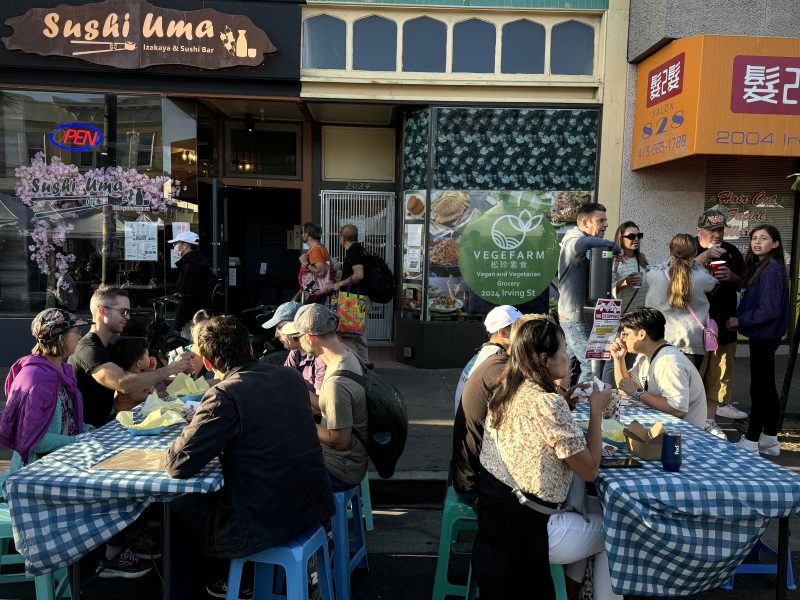 People are seated at outdoor tables eating and talking outside a row of storefronts, including a sushi bar and other restaurants, on a sunny day.
