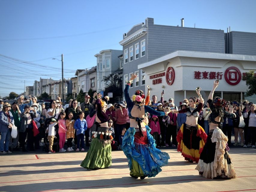 A group of dancers in colorful costumes perform outdoors on a street, surrounded by a crowd of onlookers and residential buildings in the background.