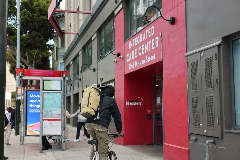 A person with a large backpack rides a bicycle past the entrance of the Integrated Care Center at 1563 Mission Street, near a covered bus stop and street map.
