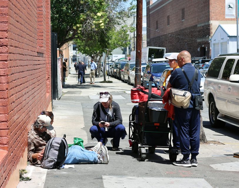 A group of people with carts talk to a man sitting on the sidewalk against a brick wall; another person kneels nearby using a phone.