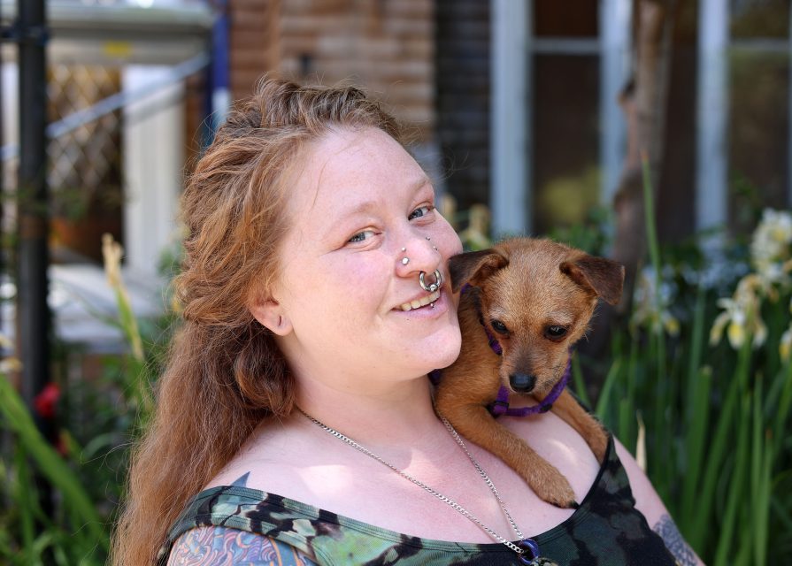 A person with long hair and facial piercings smiles while holding a small brown dog on their shoulder, standing outside near plants and a house.