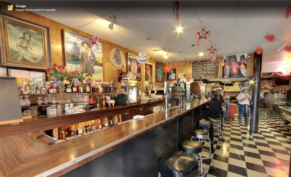 A vintage-style bar with a long counter, bar stools, checkered floor, wall art, and hanging heart decorations. Two people stand in the background near the bar area.