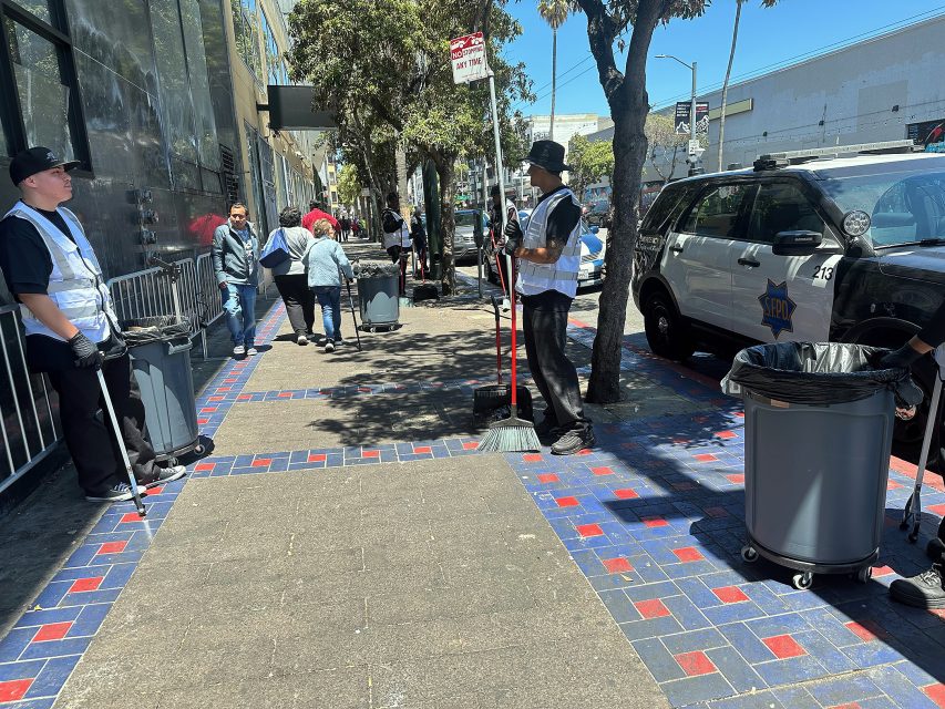 Several people clean a city sidewalk with brooms and trash bins, while pedestrians walk by and a police vehicle is parked nearby.