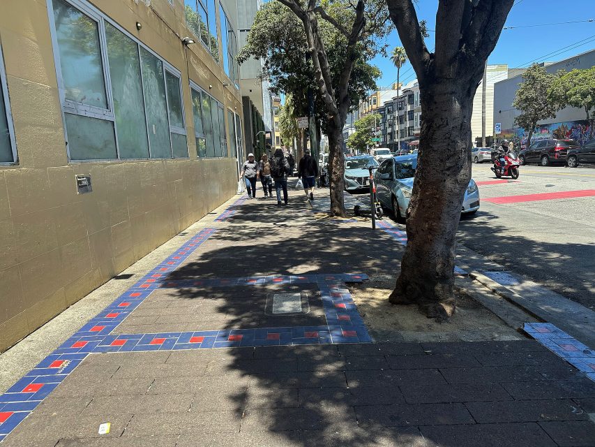 A city sidewalk with trees casting shadows, bordered by a blue and red tiled edge; pedestrians walk ahead, and parked cars line the street.