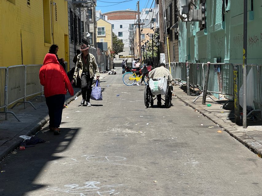 People walk and push carts down a city alley lined with barricades, graffiti, and scattered litter on a sunny day.