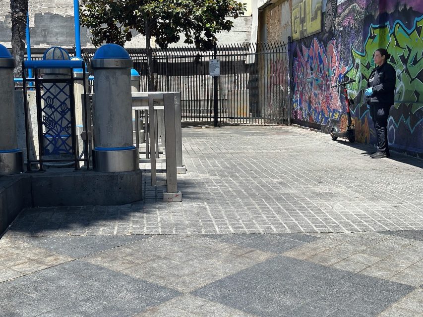 A person stands near a wall covered in colorful graffiti, next to a fenced area with metal benches and blue-topped structures in an urban outdoor setting.