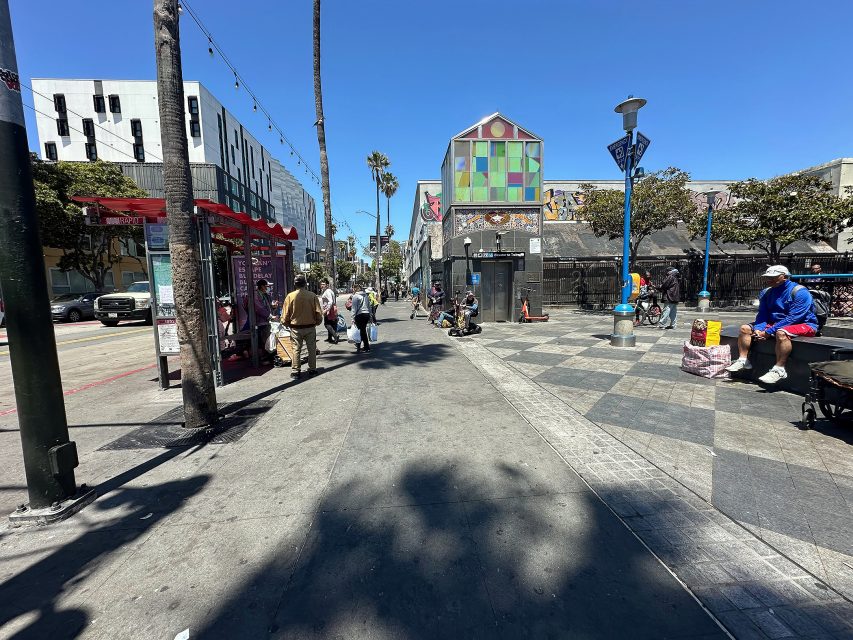 People walking and sitting along a sunny, palm-lined sidewalk with colorful buildings, street art, and bikes in the background.