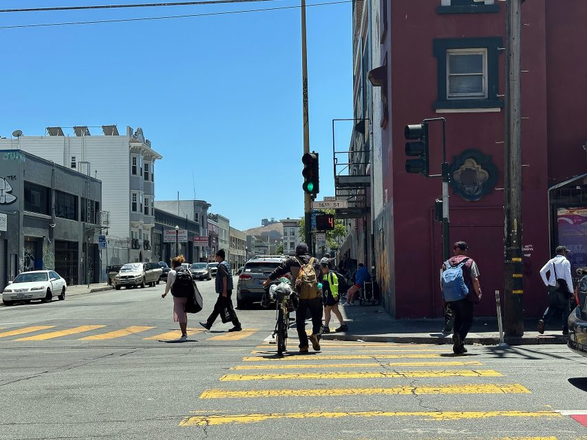People walk across a city street at a crosswalk on a sunny day, with cars parked along the curb and buildings lining both sides of the intersection.
