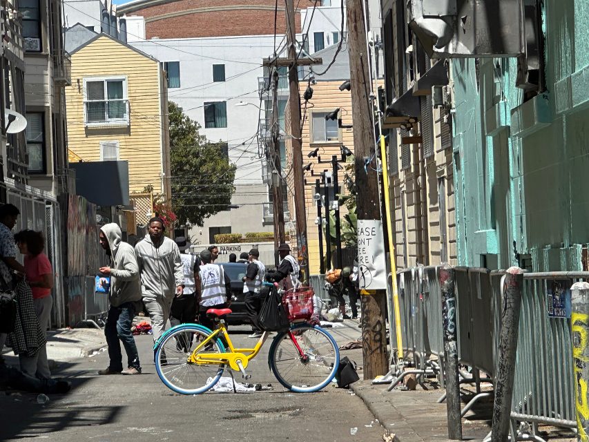 A group of people stand and walk in an urban alleyway. A yellow bicycle is parked in the foreground, with buildings and utility poles in the background.