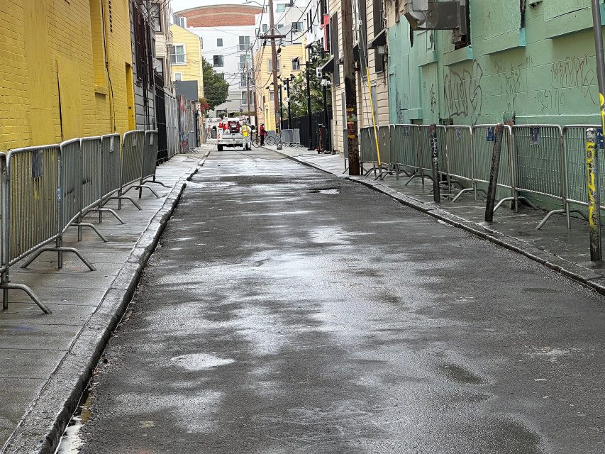 A wet, empty alley lined with metal barricades, yellow and green graffiti-covered walls, and a few people visible in the distance.