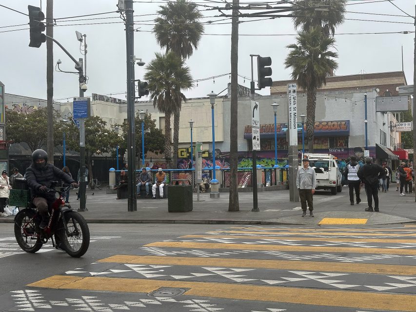 A person rides an electric bike through a crosswalk; pedestrians stand and walk near a graffiti-covered building with palm trees in the background on an overcast day.