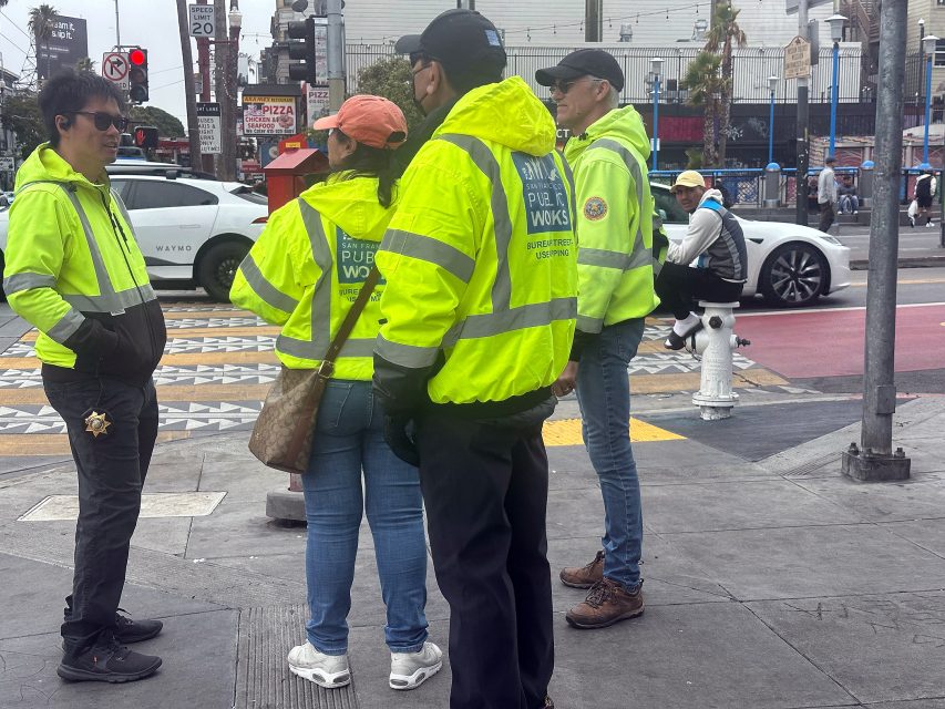 A group of people wearing bright yellow Public Works jackets stand and talk on a city sidewalk near a crosswalk.