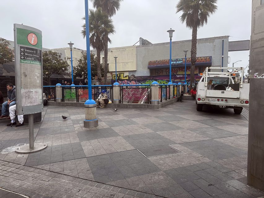 A city plaza with a few people sitting on benches, a parked utility truck, palm trees, and colorful graffiti on the background wall.