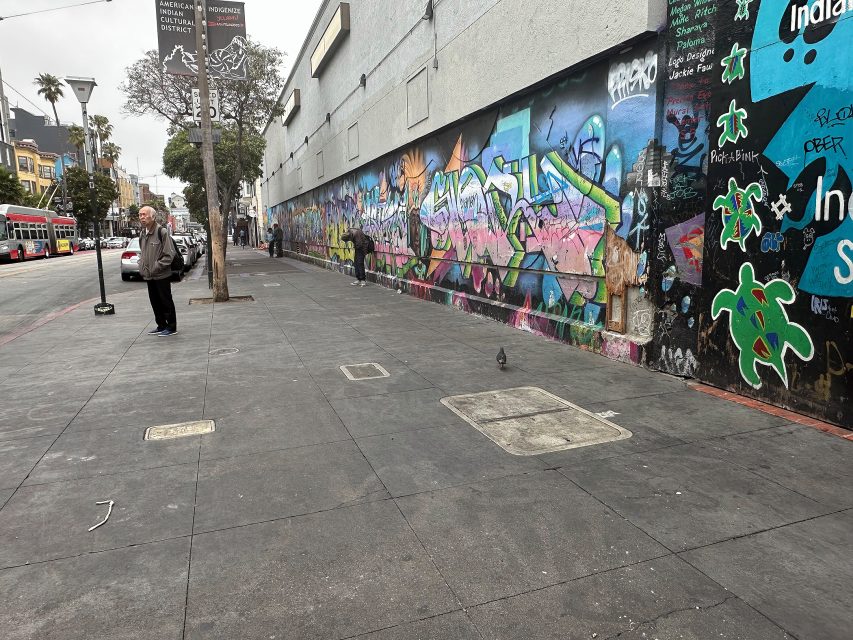 A man stands on a city sidewalk next to a building with colorful graffiti murals; a pigeon is on the pavement nearby.