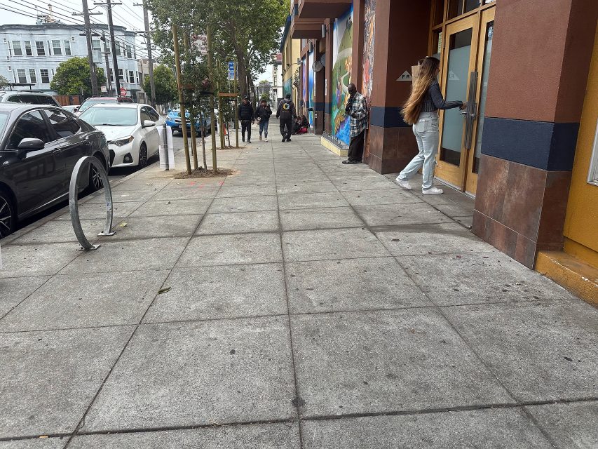 People walk along a city sidewalk lined with parked cars and storefronts; one person opens a door while others are further down the street.
