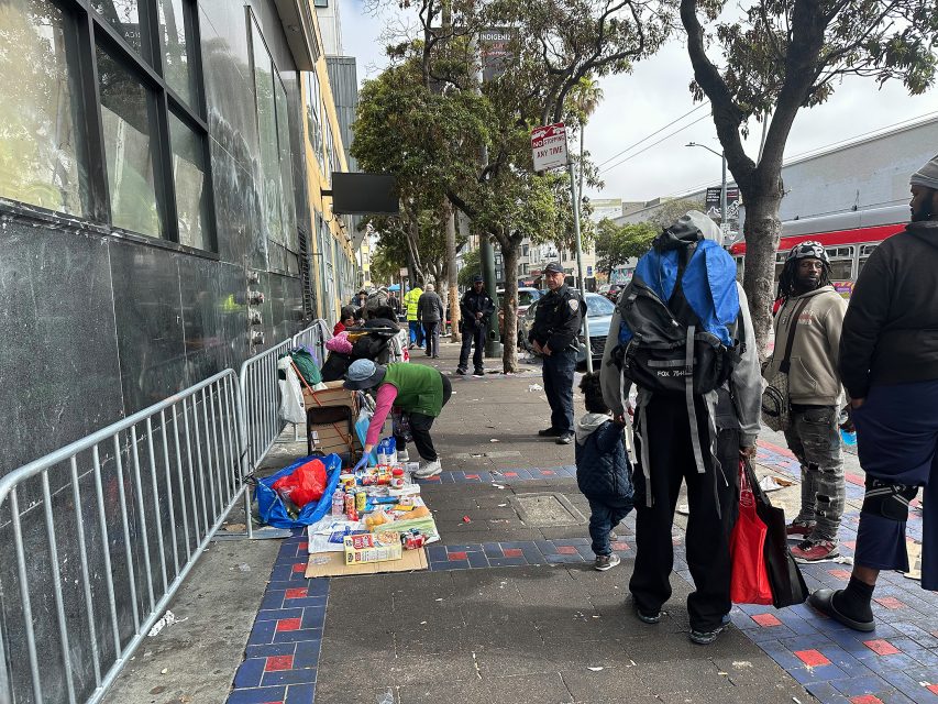 People gather on a city sidewalk with items laid out for sale; police officers stand nearby and trees line the street.