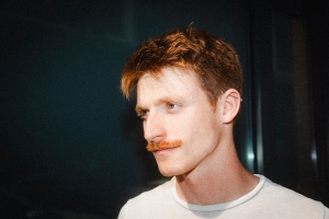 A young man with light skin, short reddish-brown hair, and a mustache looks off to the side. He is wearing a white shirt and stands against a dark background.