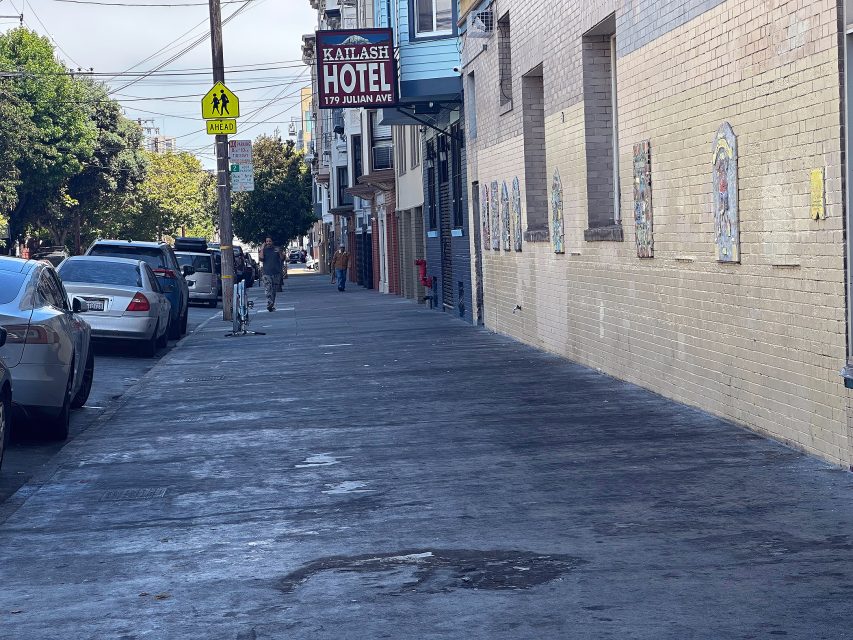 A city sidewalk with parked cars, a pedestrian crossing sign, and the Kailash Hotel sign above 179 Julian Ave. Several people are visible in the background.