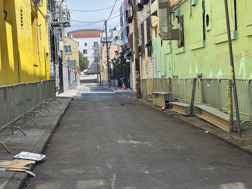 A narrow city alley with metal barricades on both sides, graffiti on the walls, and cardboard debris on the ground. Buildings border the alley, and the street is empty.