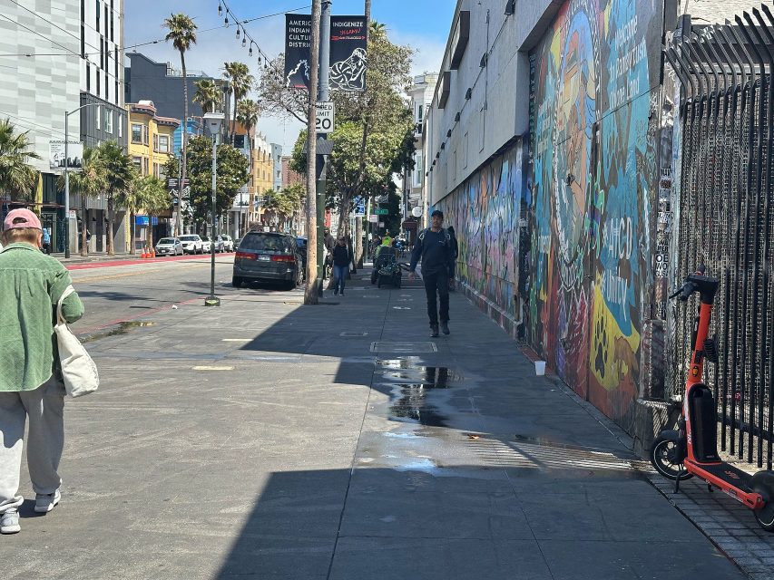 People walk along a sunny city sidewalk lined with murals, parked cars, and a parked scooter; palm trees and buildings are visible in the background.