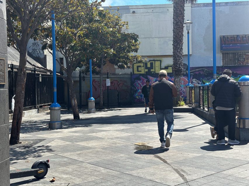 Several people walk through a sunny urban area with graffiti on the walls, trees, blue lamp posts, and a fallen electric scooter on the ground.