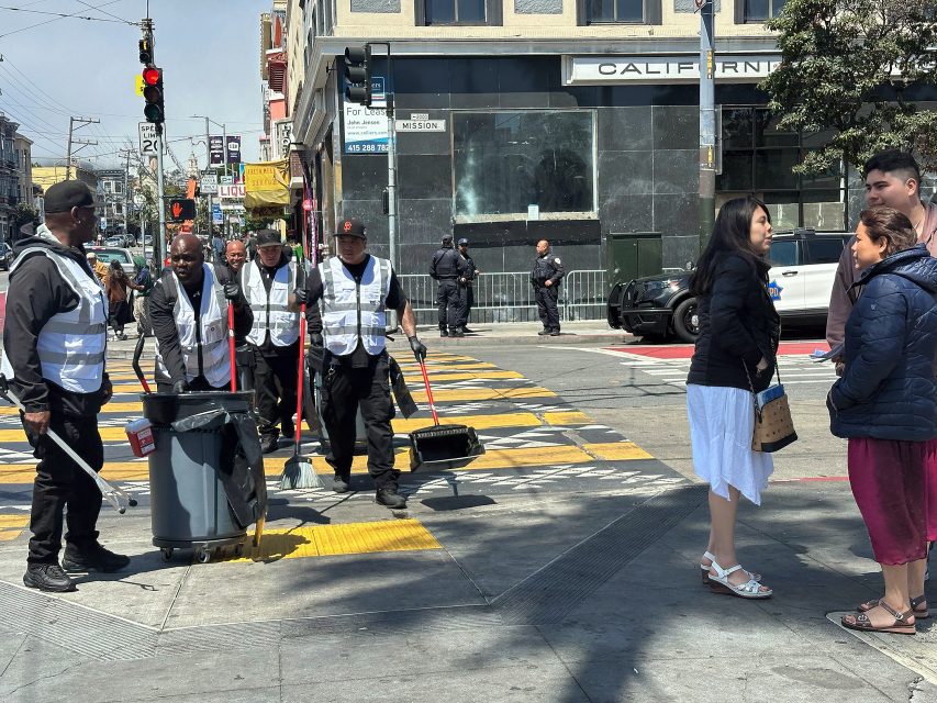 A group of city workers with cleaning equipment stand at a crosswalk while pedestrians talk on a sunny urban street.