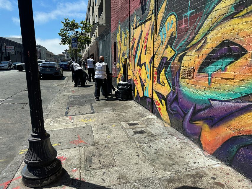 Several people in uniforms clean a sidewalk next to a colorful graffiti-covered brick wall on a sunny day in an urban area.