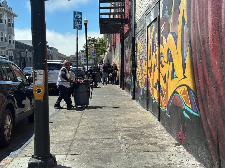 A man pushes a cart down a city sidewalk lined with parked cars and buildings, one of which has a large colorful graffiti mural. Several people are gathered in the distance.