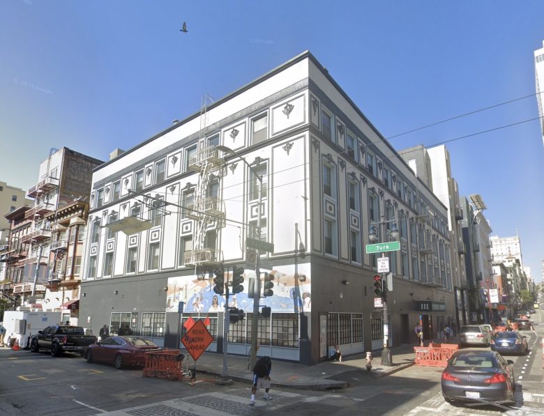 A three-story white and gray building with geometric designs on its facade, located at a street corner with traffic signs, cars, and pedestrians nearby.