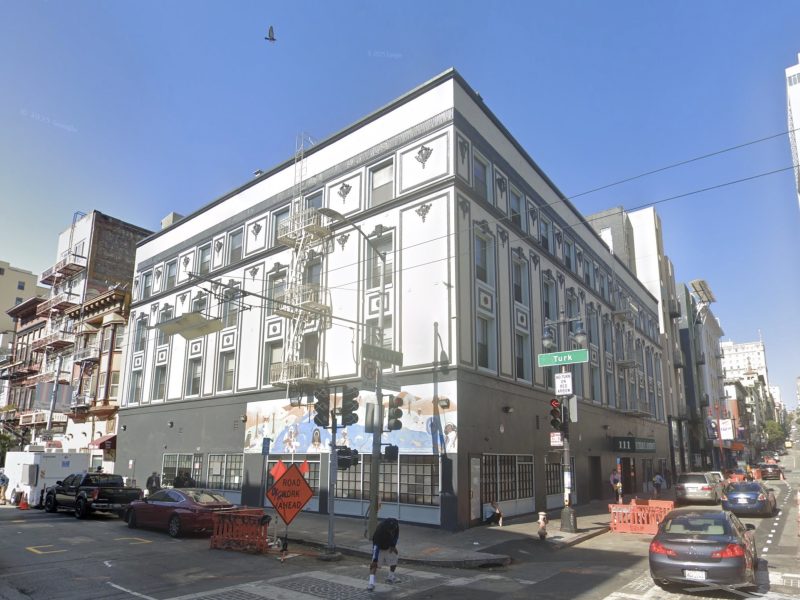 A three-story white and gray building with geometric designs on its facade, located at a street corner with traffic signs, cars, and pedestrians nearby.