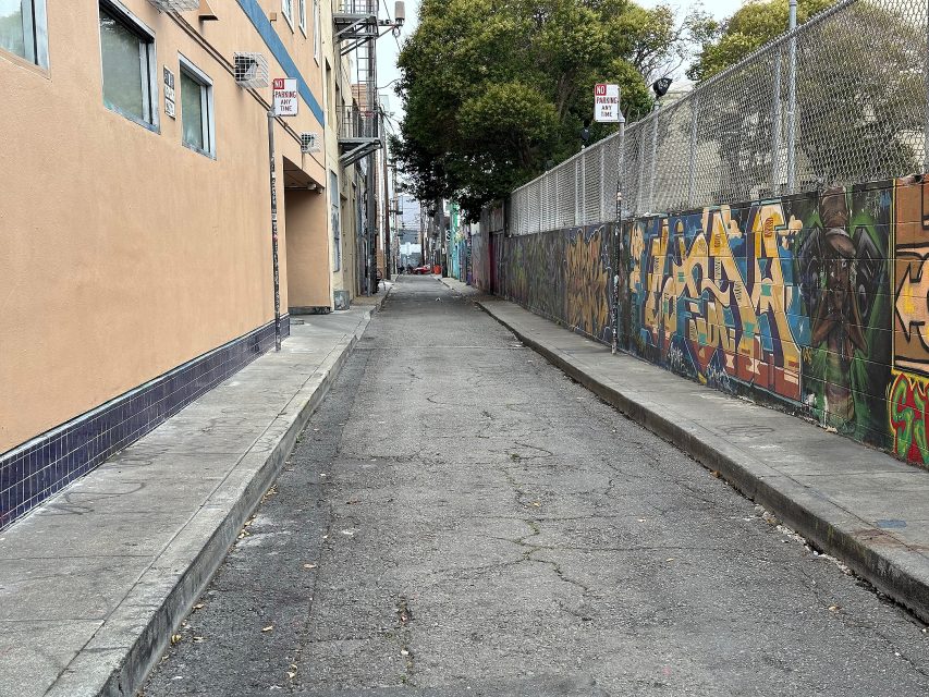 A narrow urban alley with cracked pavement, a beige building on the left, and a graffiti-covered wall with a chain-link fence on the right.