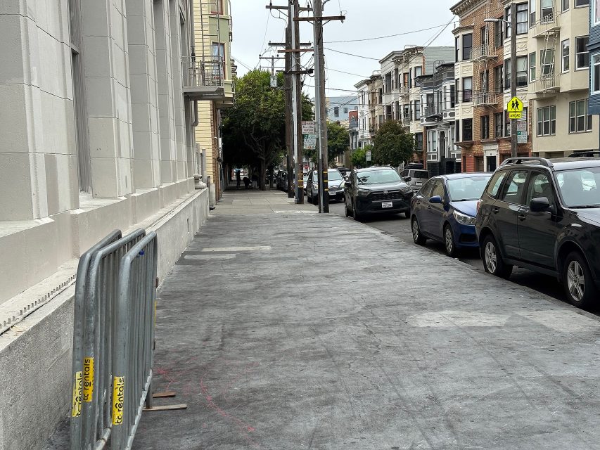Wide city sidewalk with temporary barricades on the left, parked cars on the right, and apartments lining the street in the background.