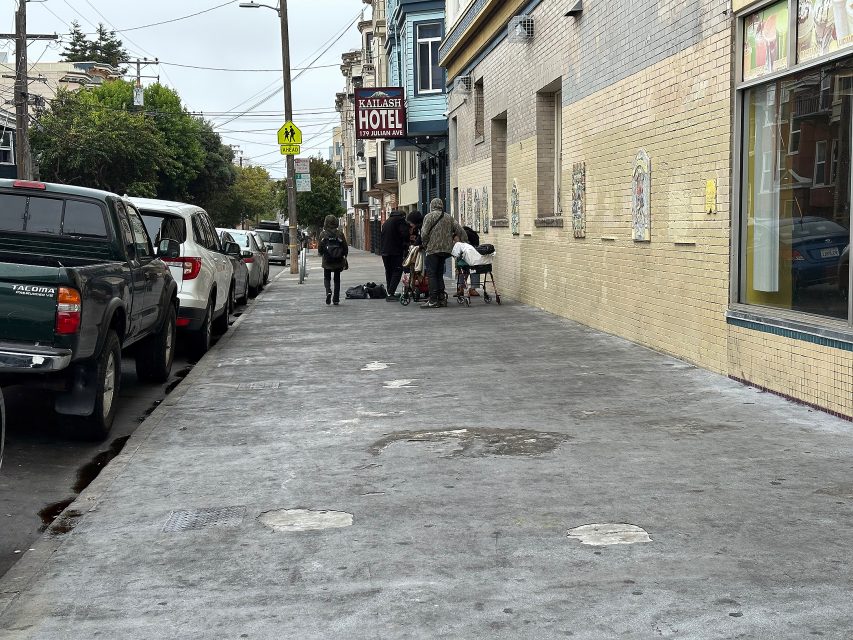 A group of people with belongings gather near the wall of a building labeled "Royan Hotel" on a city sidewalk lined with parked cars.