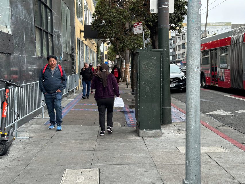 People walk on a city sidewalk next to a street with a bus and parked cars; buildings, trees, and a metal fence line the left side.
