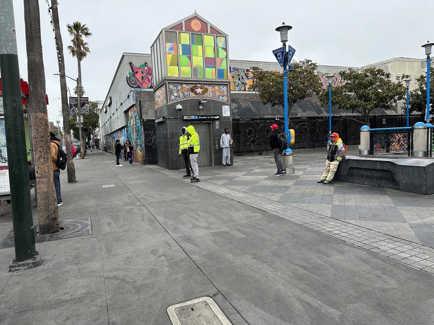 A city street scene with several people standing and sitting near a building with colorful geometric windows and graffiti on its walls.